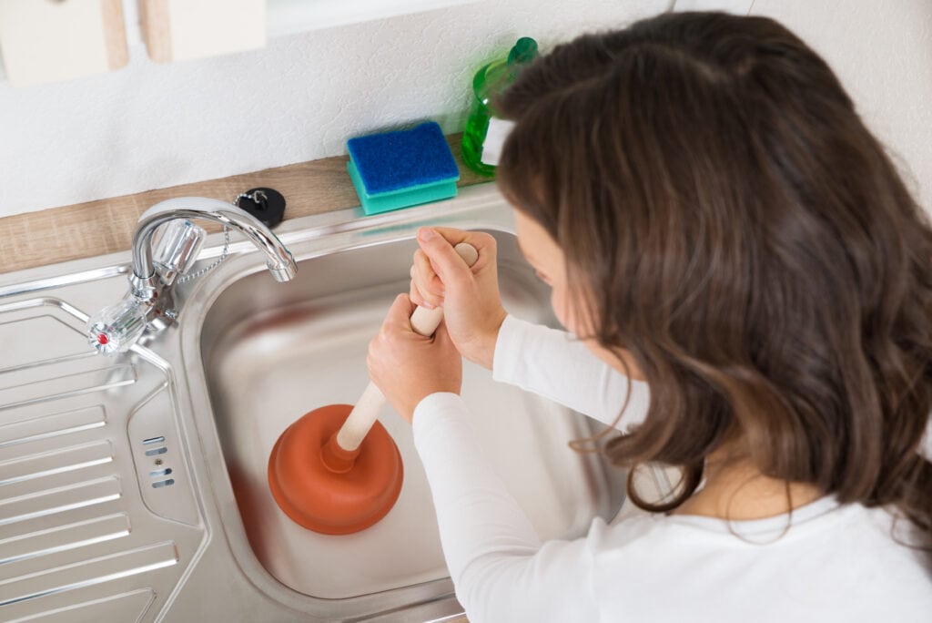 Why Is My Garbage Disposal Humming? - Happy Woman Using Plunger In Kitchen Sink At Home