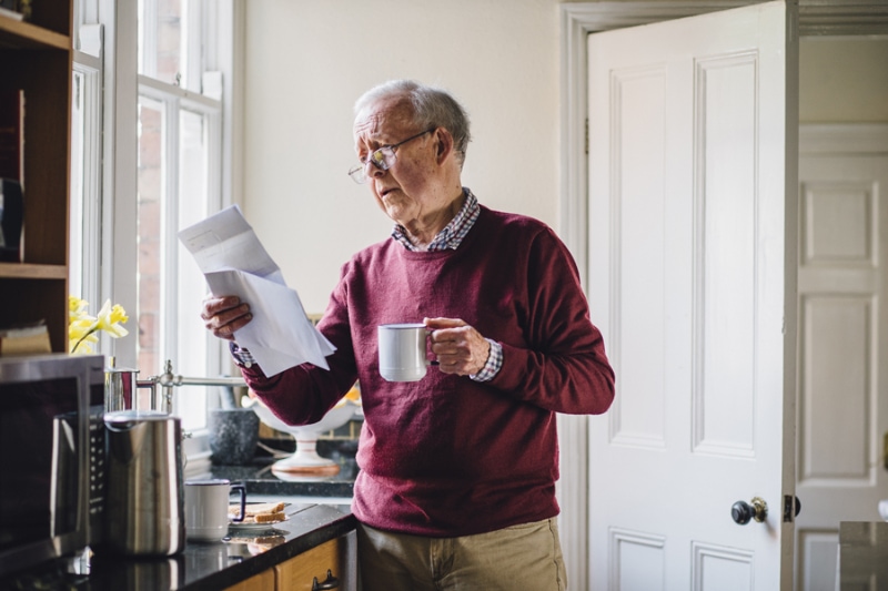 What Problems Can a Leaky Faucet Cause? -Senior man is standing in the kitchen of his home with bills in one hand and a cup of tea in the other. He has a worried expression on his face.