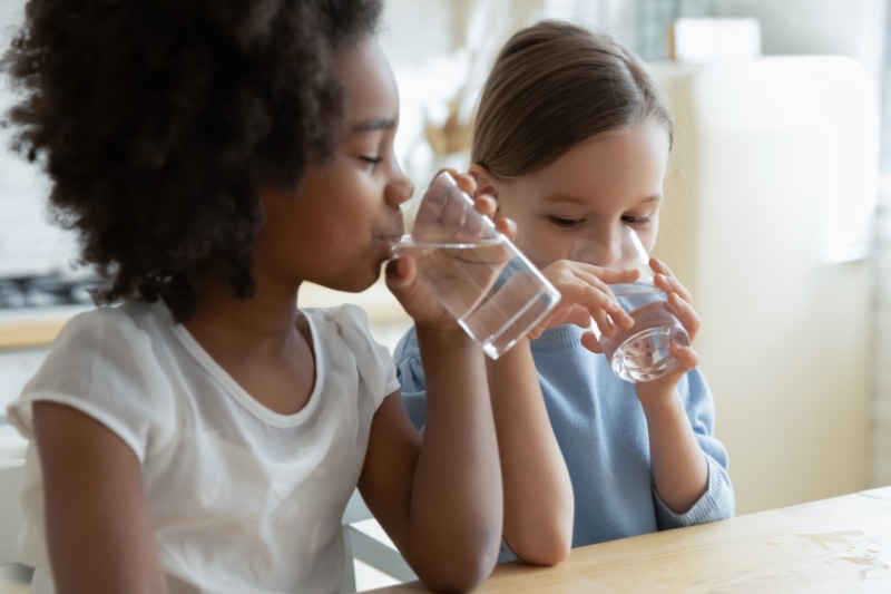 Reduce Microplastics In Your Drinking Water. Two multi racial little girls sit at table in kitchen feels thirsty drink clean still natural or mineral water close up image. Healthy life habit of kids, health benefit dehydration prevention concept.